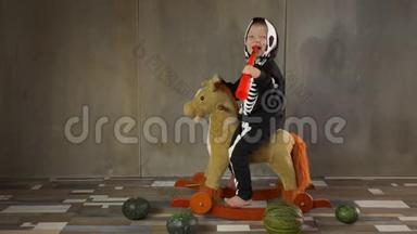 小男孩光着脚穿着骷髅服骑着摇摆玩具马。 儿童快乐庆祝万圣节，手持南瓜形手电筒